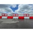 Row of alternating red and white Defender50 water-filled barriers with plastic fencing tops, installed on a large tarmac surface under cloudy skies