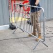 A worker in gloves and tan trousers positions a galvanised steel crowd control barrier with fixed legs on a paved surface near a temporary site enclosure.